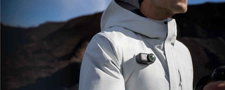 Person wearing a light gray jacket with a DJI Osmo Nano on the chest against a mountainous background. Sold by an Authorized DJI Retailer.